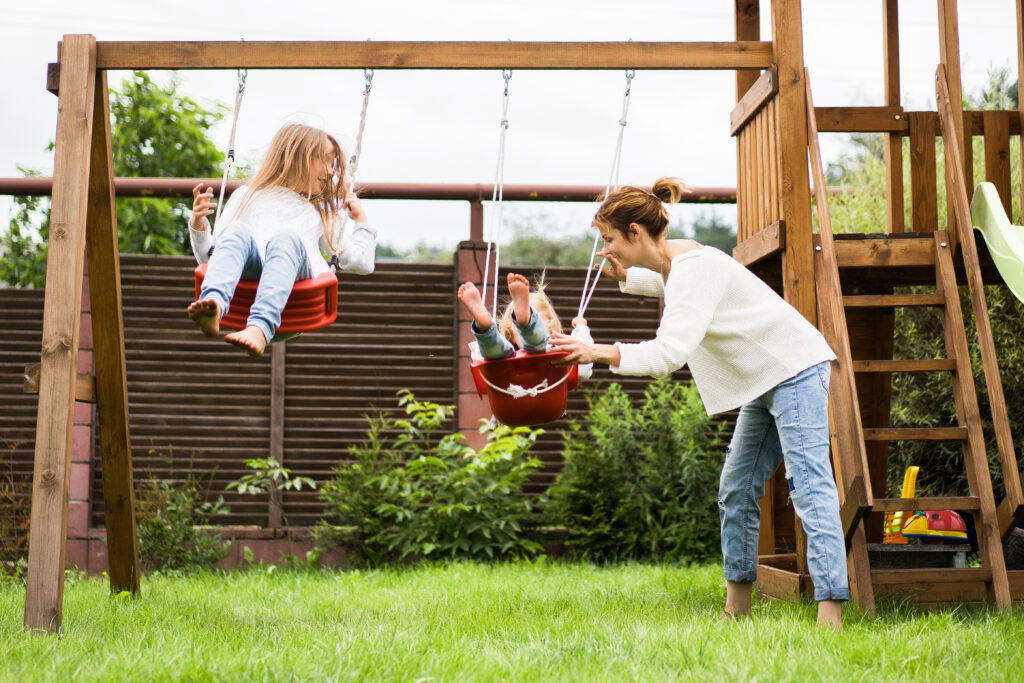 Sisters on a swing set being pushed by their mother in the garden