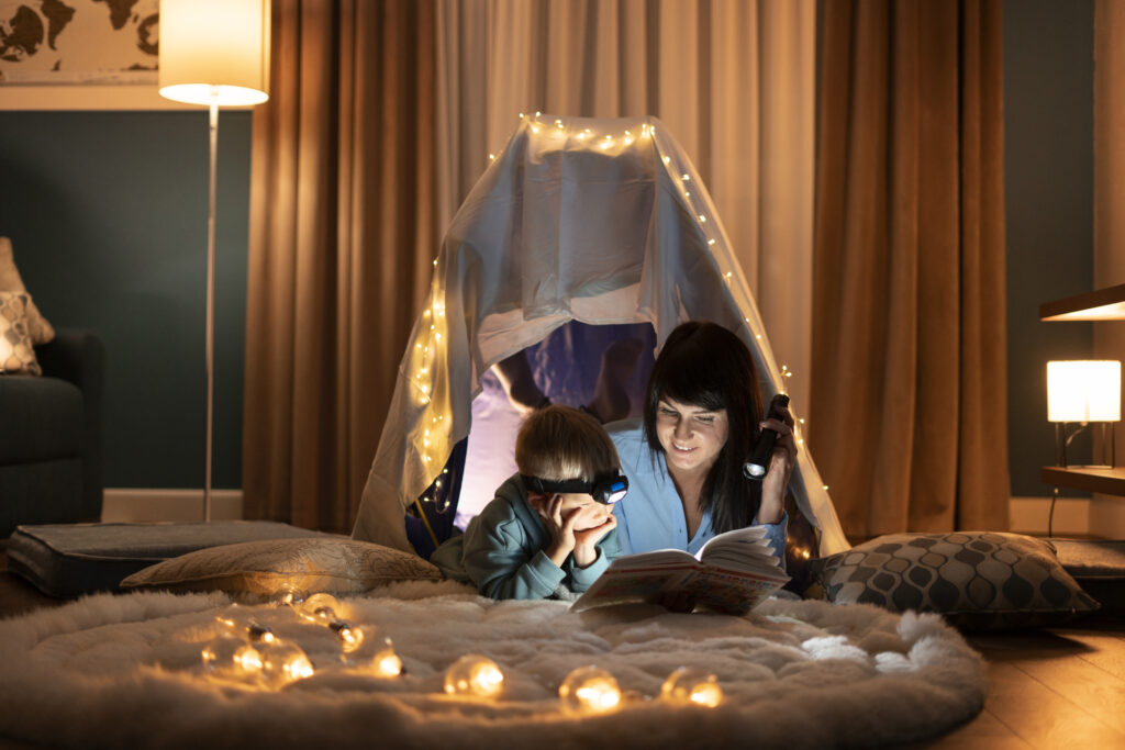 Child and mother in homemade fort with fairy lights reading a book